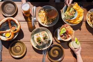 Outdoor dining table at Nelson’s, Terranea Resort in Rancho Palos Verdes, featuring pretzels, tacos, burgers, fries, guacamole, and cocktails with ocean-view ambiance.