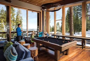 Families enjoying billiards at The Treehouse clubhouse at Northstar Resort, with large windows revealing snowy pine-covered mountains in the background