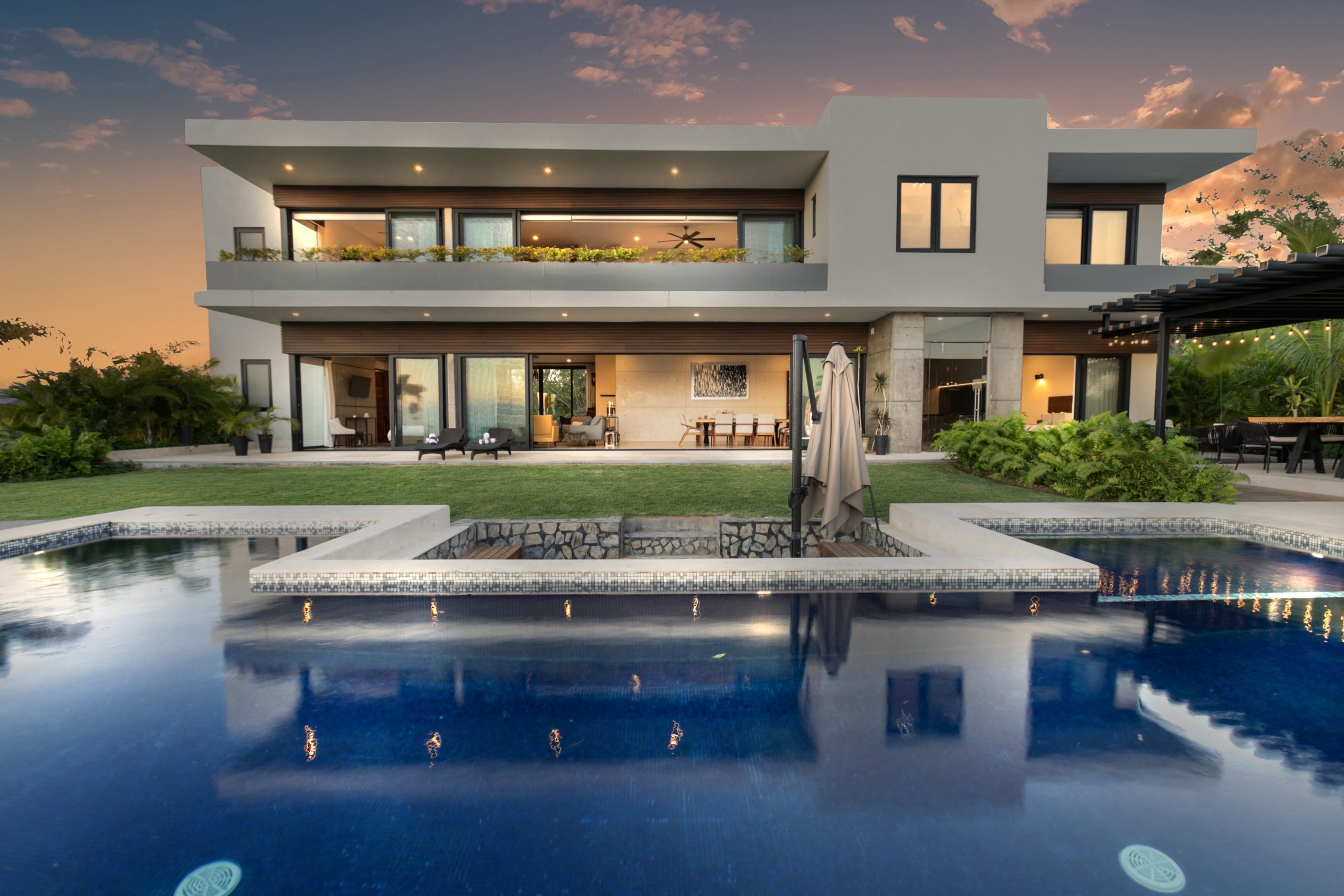 Modern two-story coastal home with white walls, large glass windows, and an infinity pool surrounded by lush greenery under a clear blue sky
