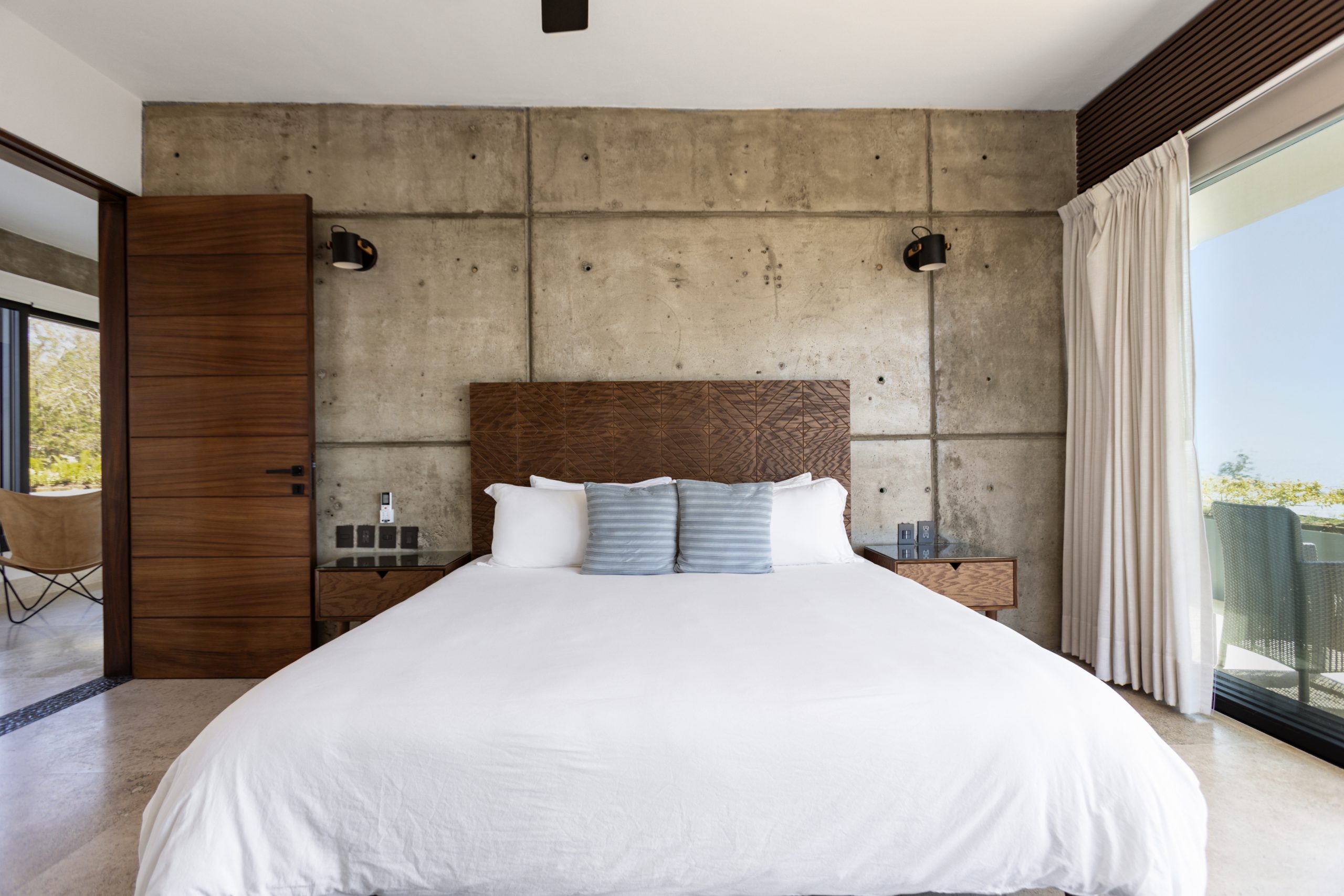 edroom with panoramic ocean views through large corner windows, light-colored curtains, and a cozy chair with a blanket.