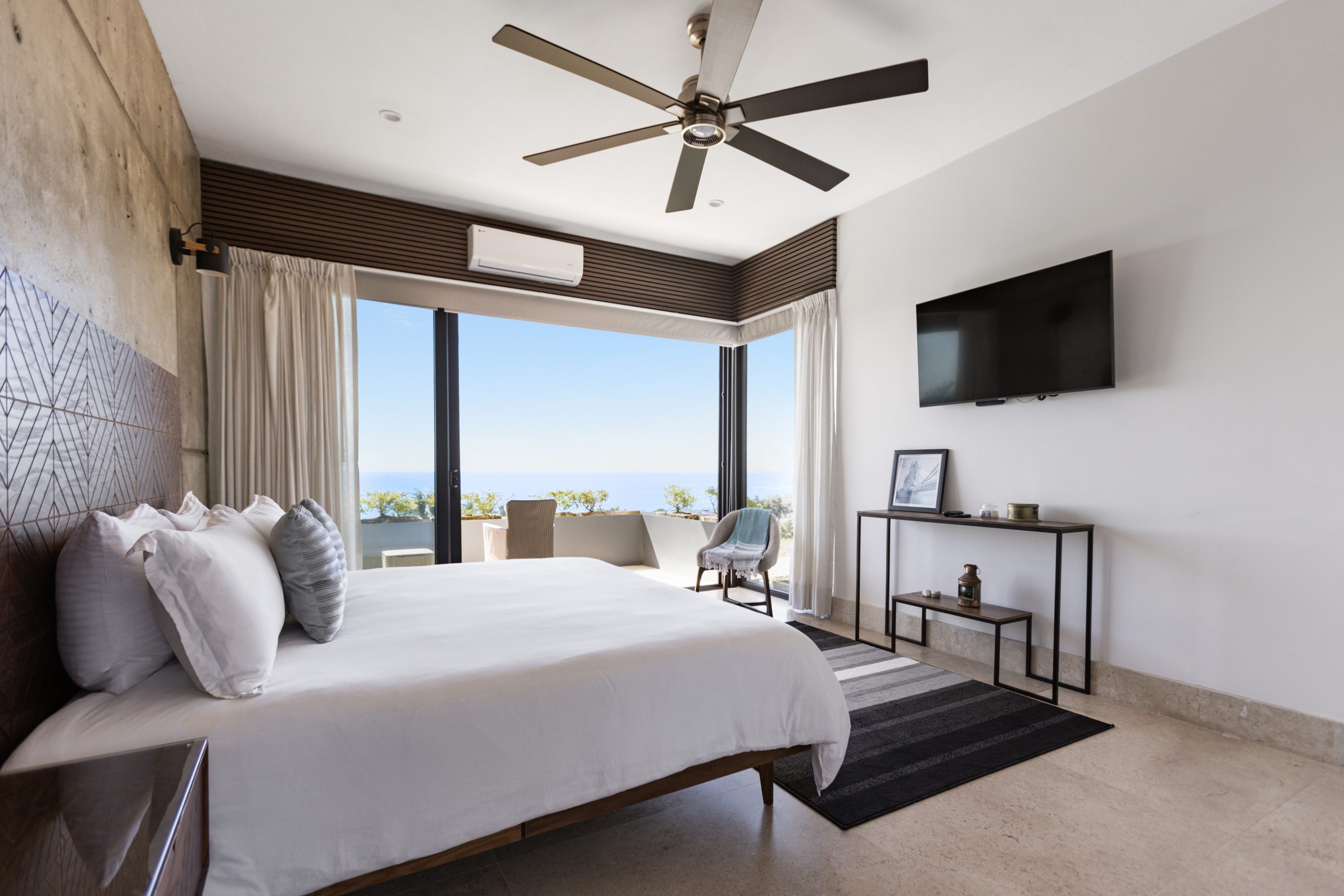 edroom with panoramic ocean views through large corner windows, light-colored curtains, and a cozy chair with a blanket.