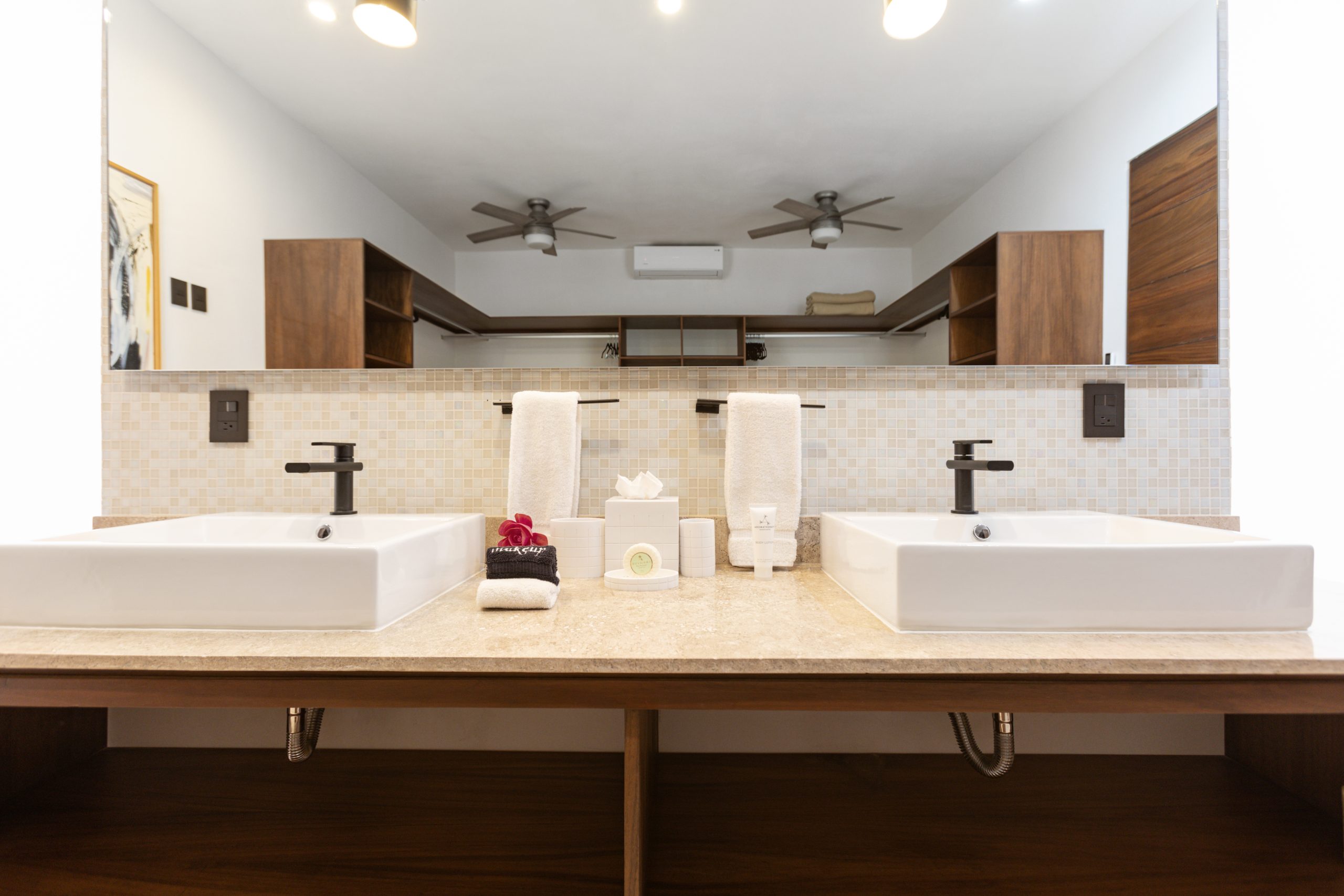 Modern bathroom with double sinks, large mirror, beige stone countertop, and wooden shelving reflected in the background.