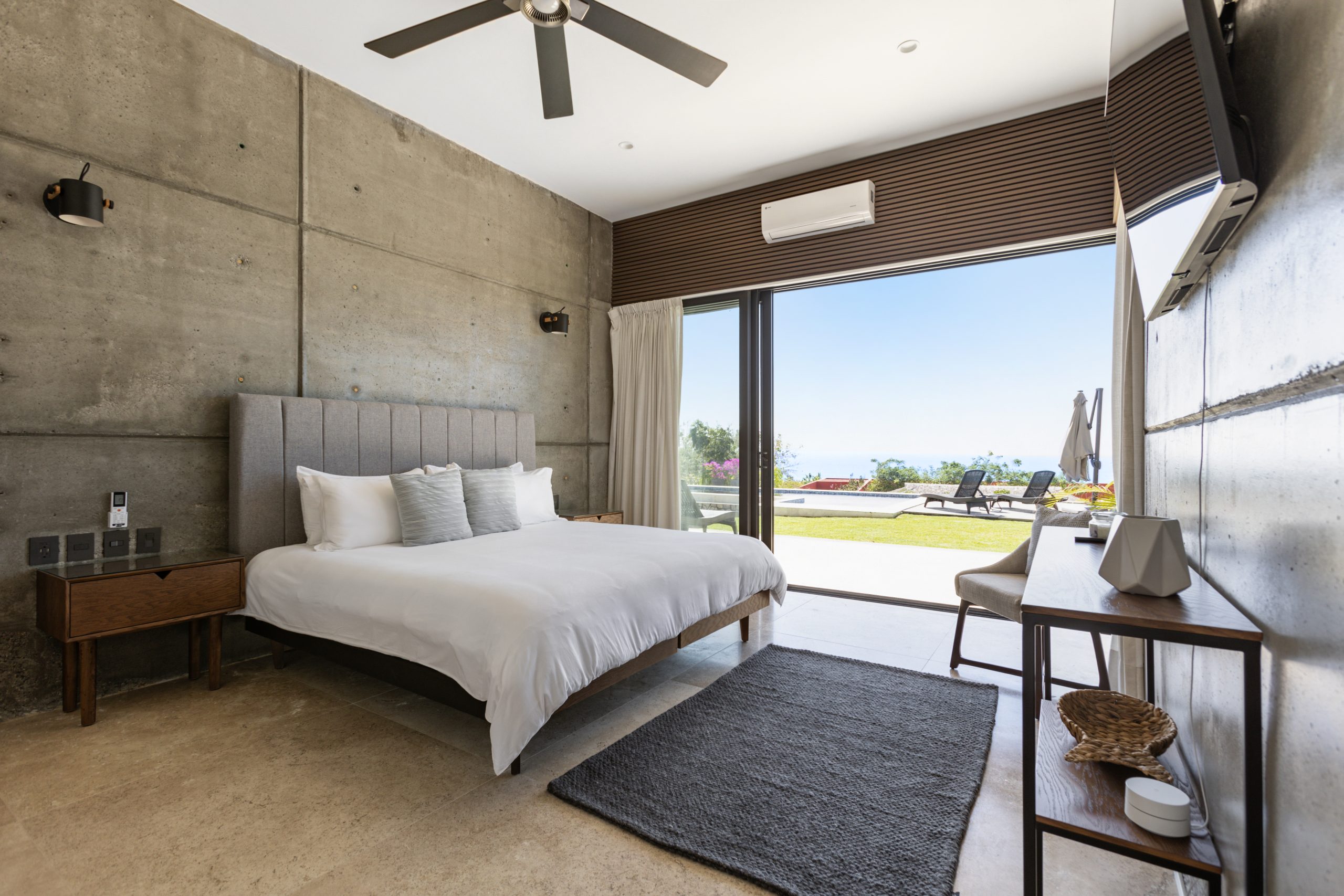 edroom with panoramic ocean views through large corner windows, light-colored curtains, and a cozy chair with a blanket.