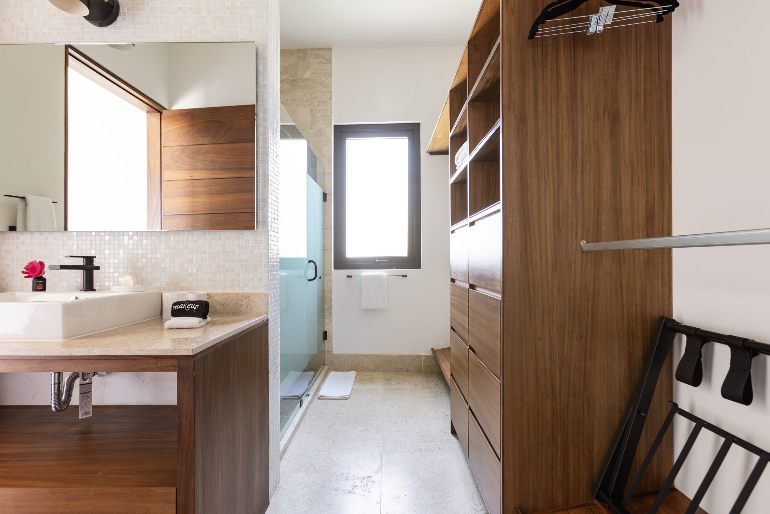 Modern bathroom with beige stone countertop, rectangular sink, and adjacent wooden walk-in closet featuring open shelving and hanging racks.