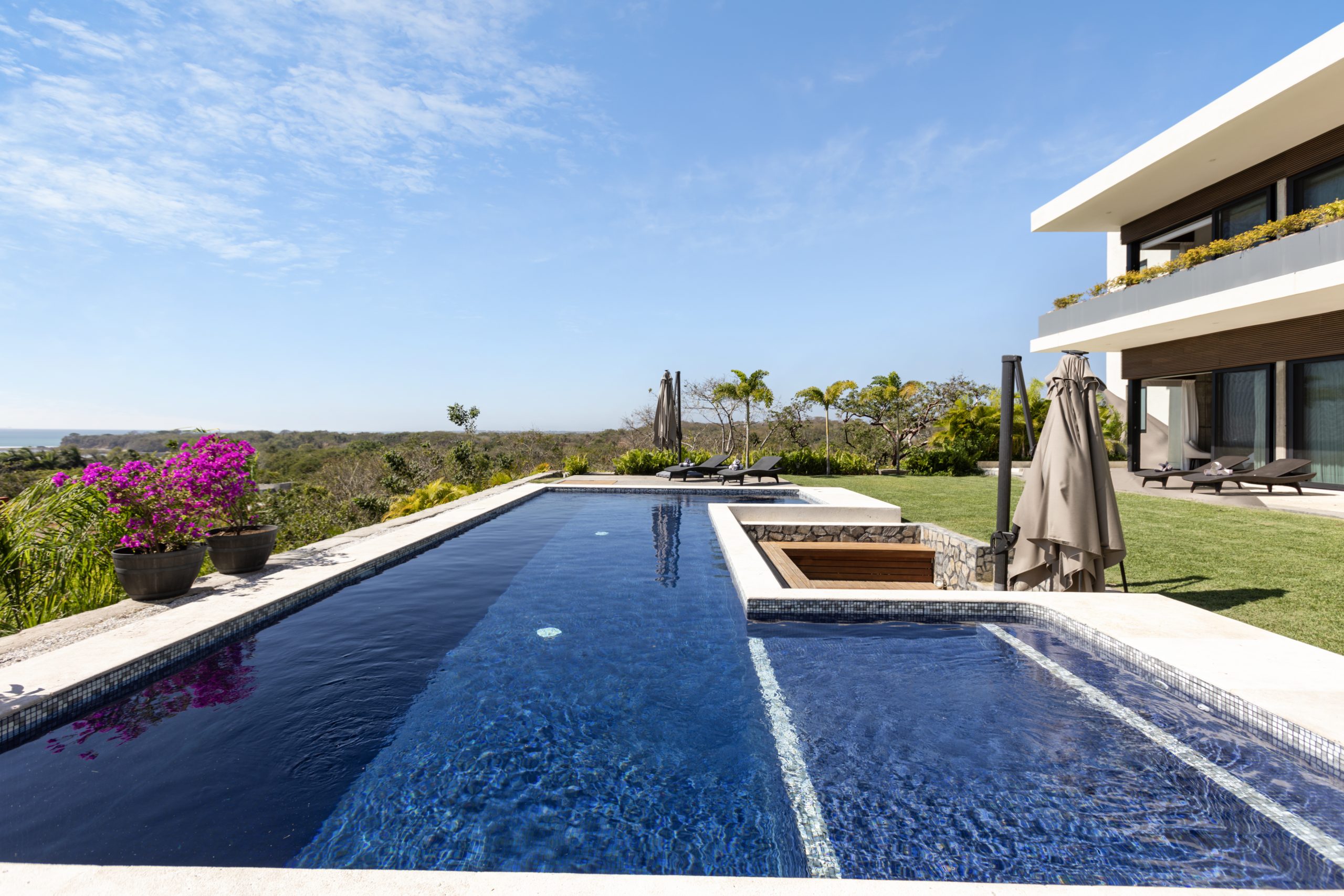 Modern two-story coastal home with white walls, large glass windows, and an infinity pool surrounded by lush greenery under a clear blue sky