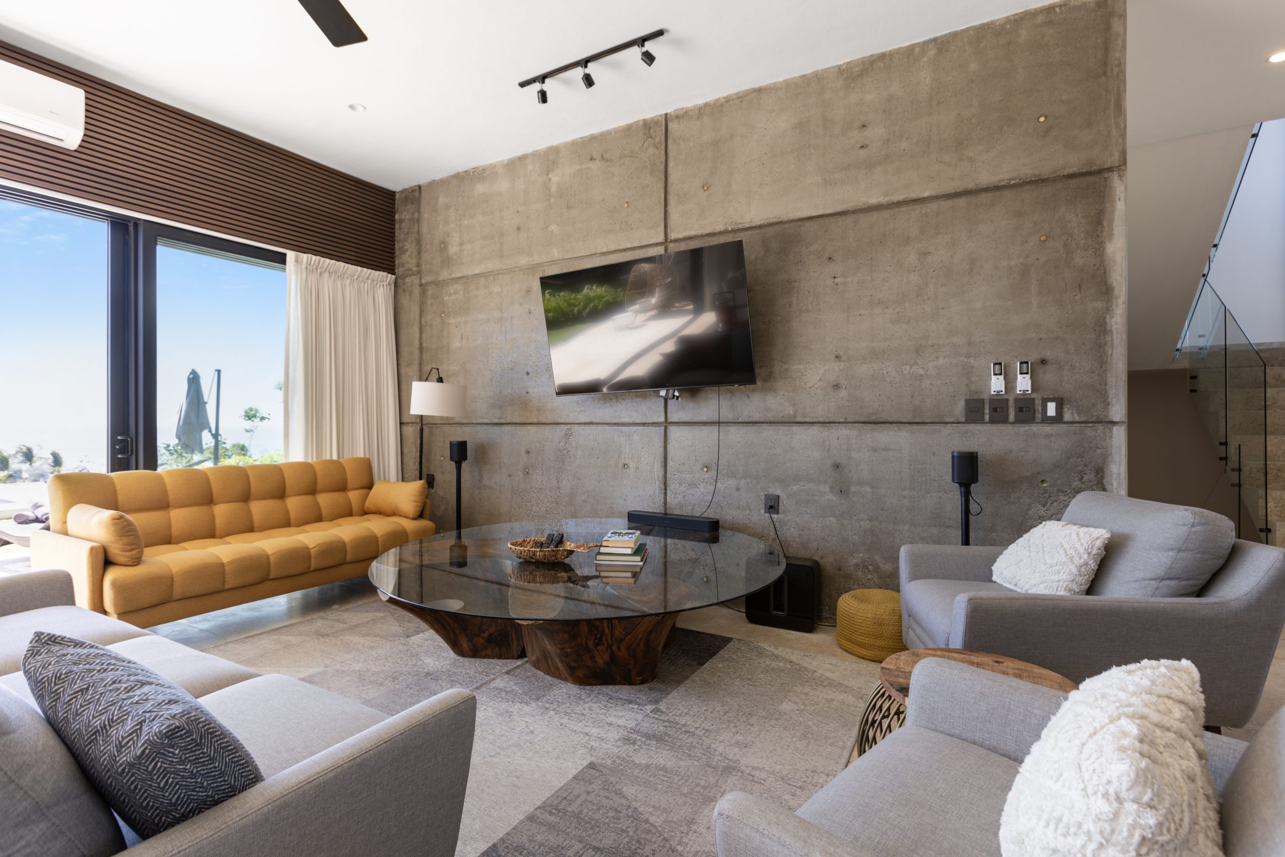 Modern living room with concrete accent wall, large TV, yellow sofa, gray armchairs, and glass doors opening to a pool view.