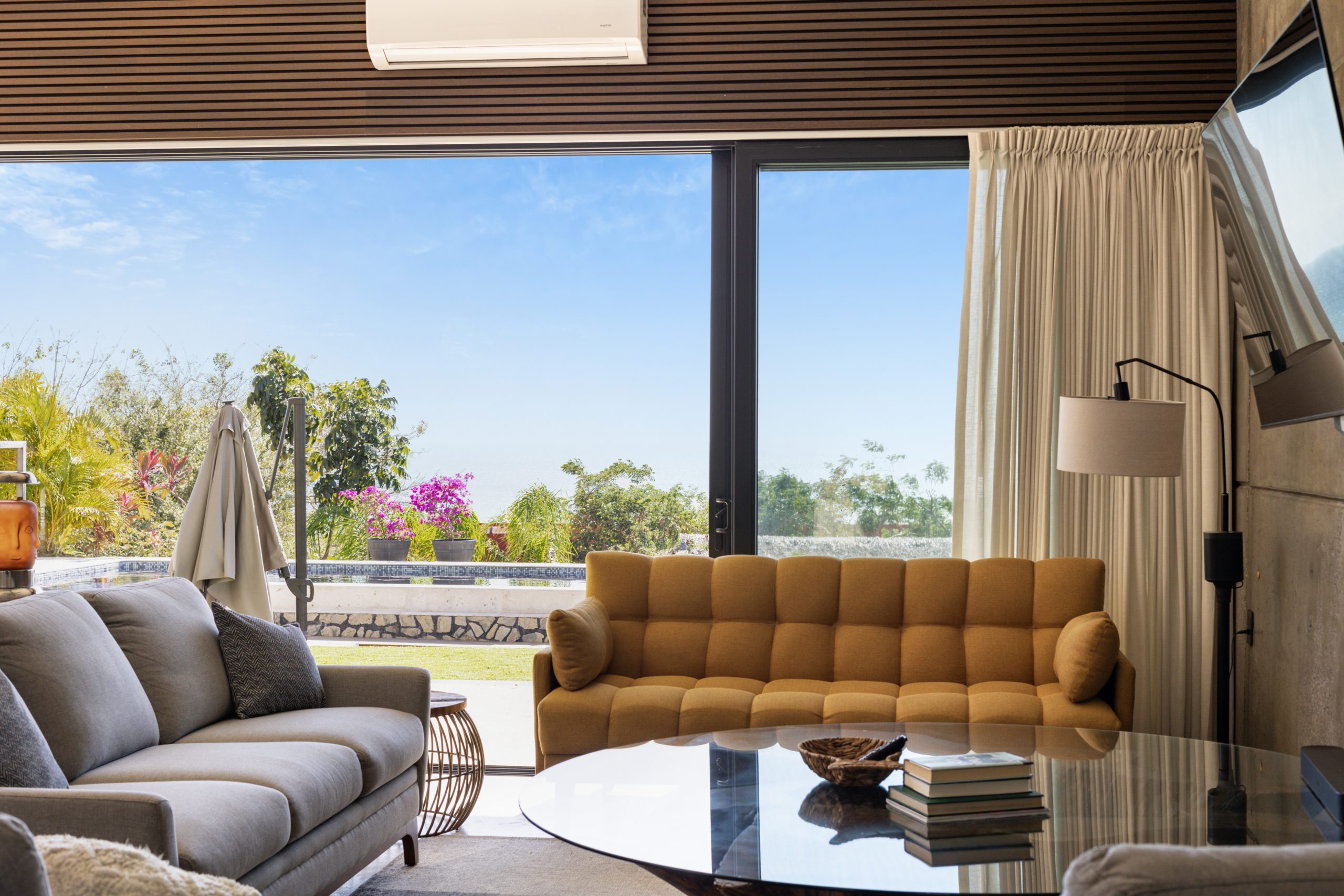 Modern living room with concrete accent wall, large TV, yellow sofa, gray armchairs, and glass doors opening to a pool view.