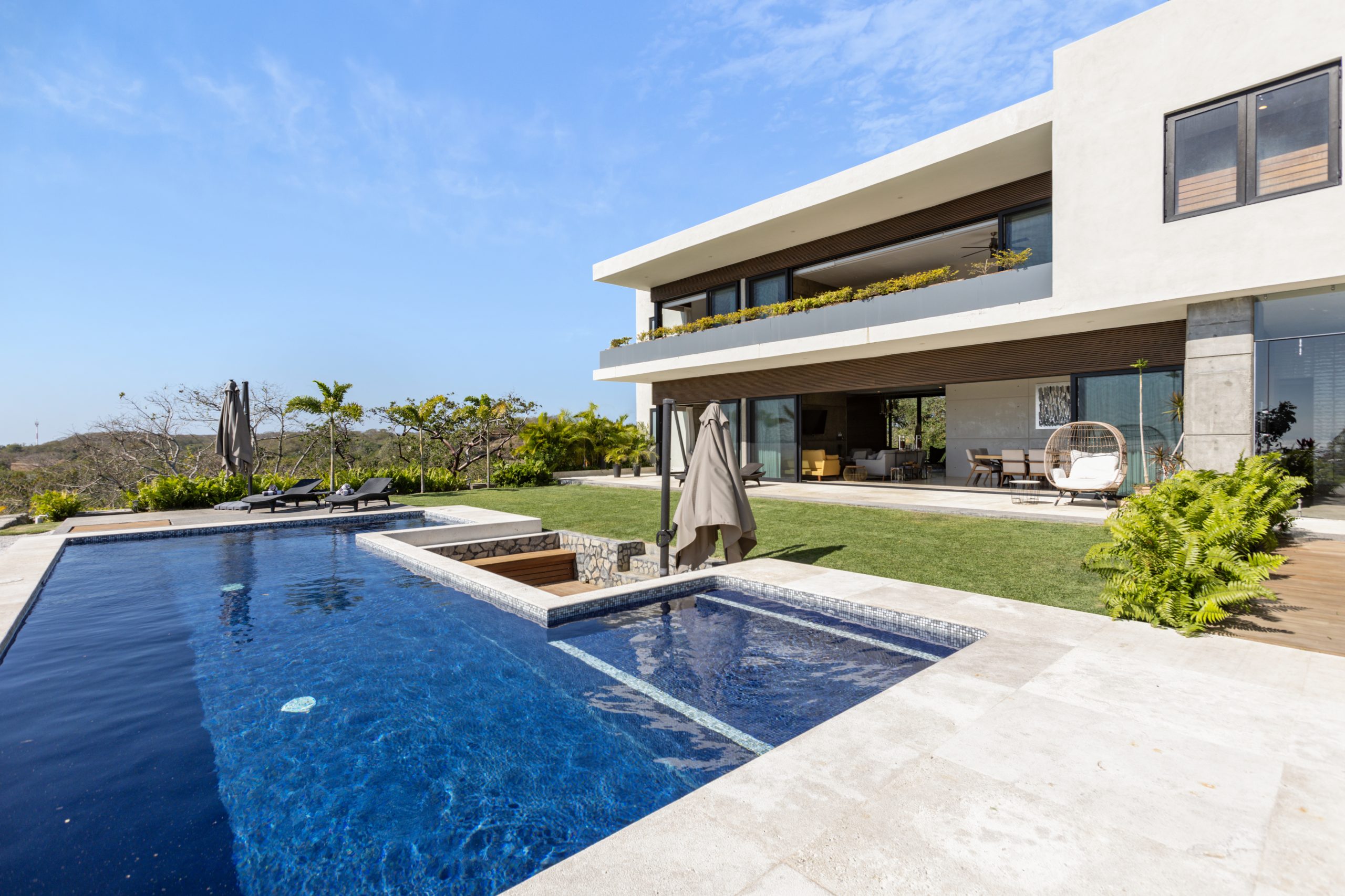 Modern two-story coastal home with white walls, large glass windows, and an infinity pool surrounded by lush greenery under a clear blue sky