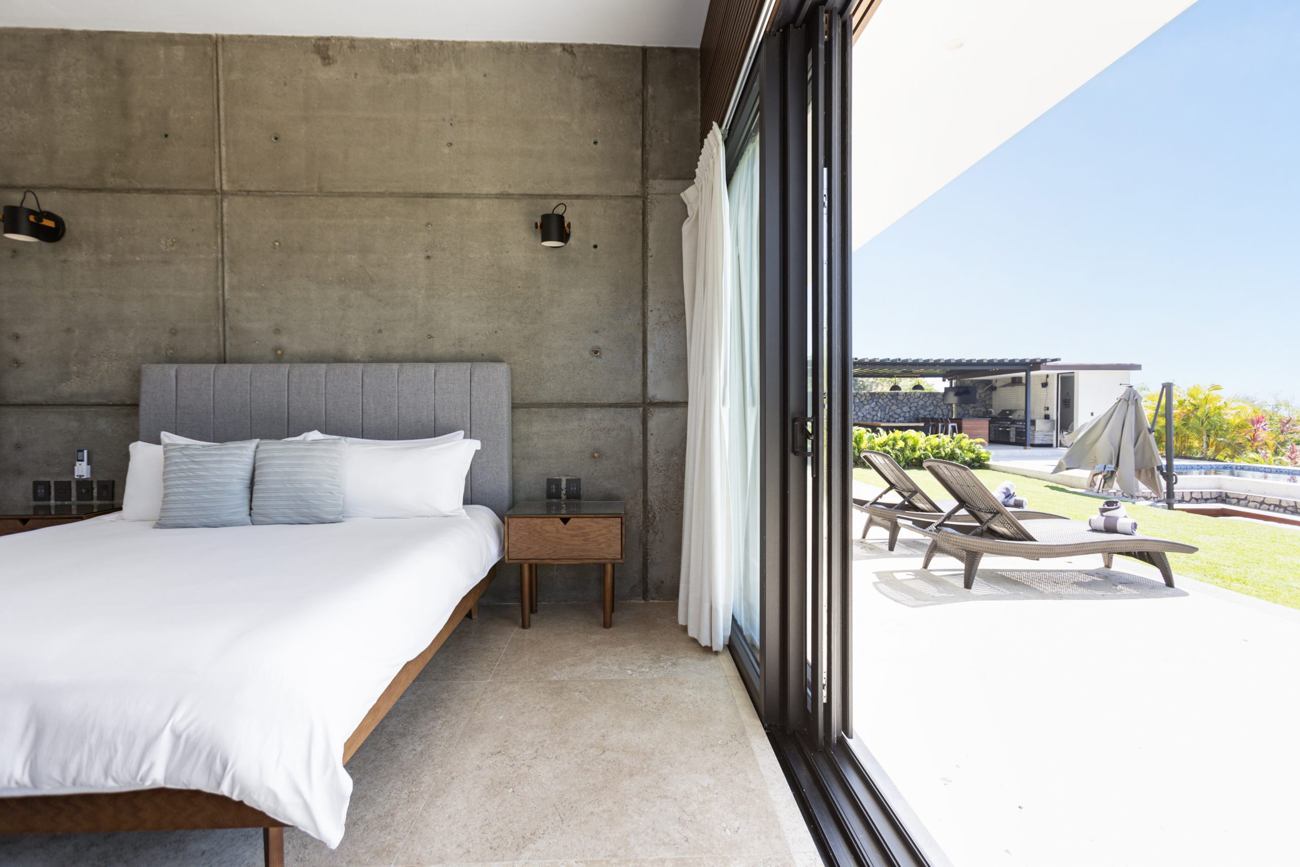 edroom with panoramic ocean views through large corner windows, light-colored curtains, and a cozy chair with a blanket.