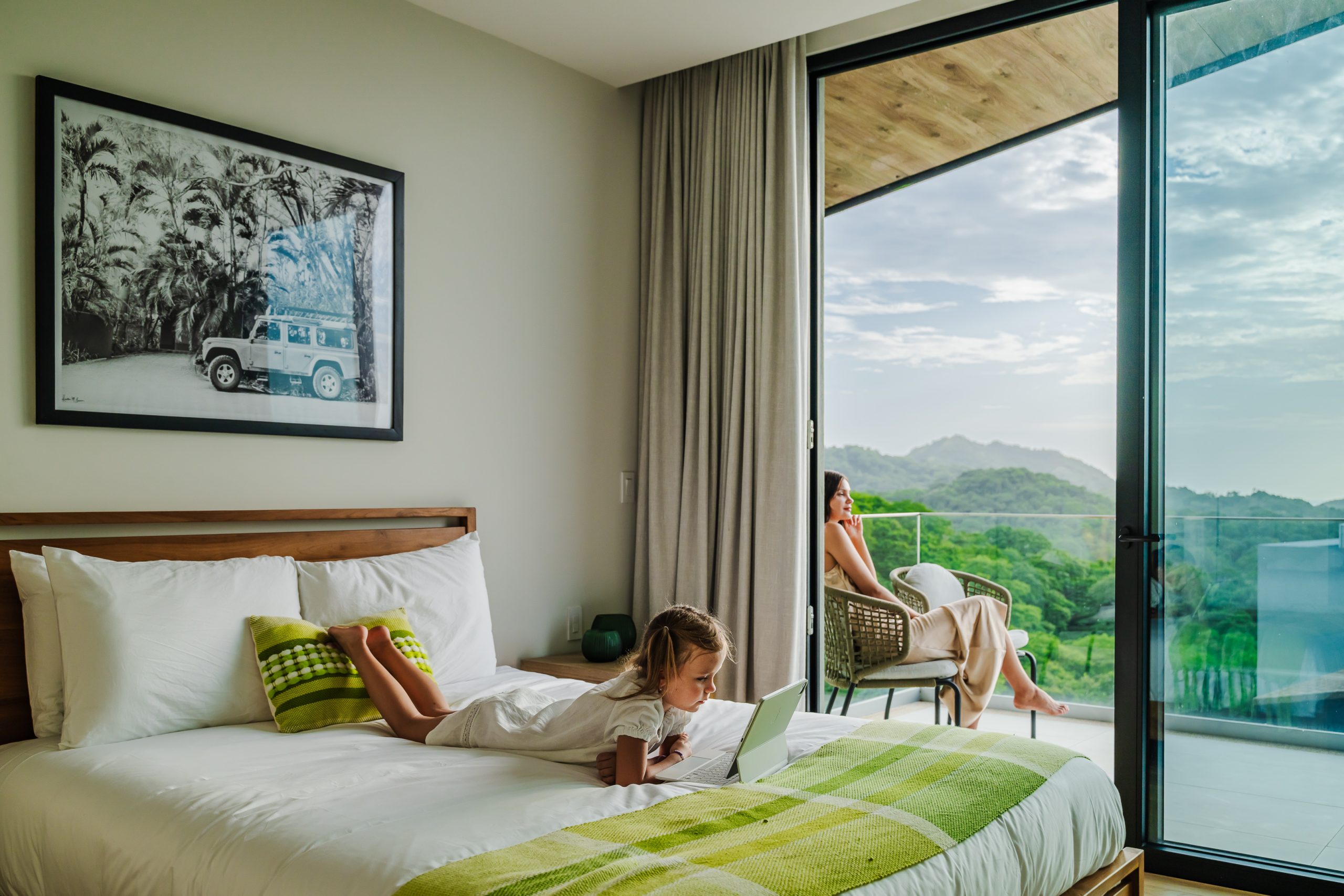 Woman relaxing on the terrace while her child lounges on the bed playing on an iPad inside the Reserva Conchal penthouse, blending comfort and family leisure in a luxury Costa Rican setting.