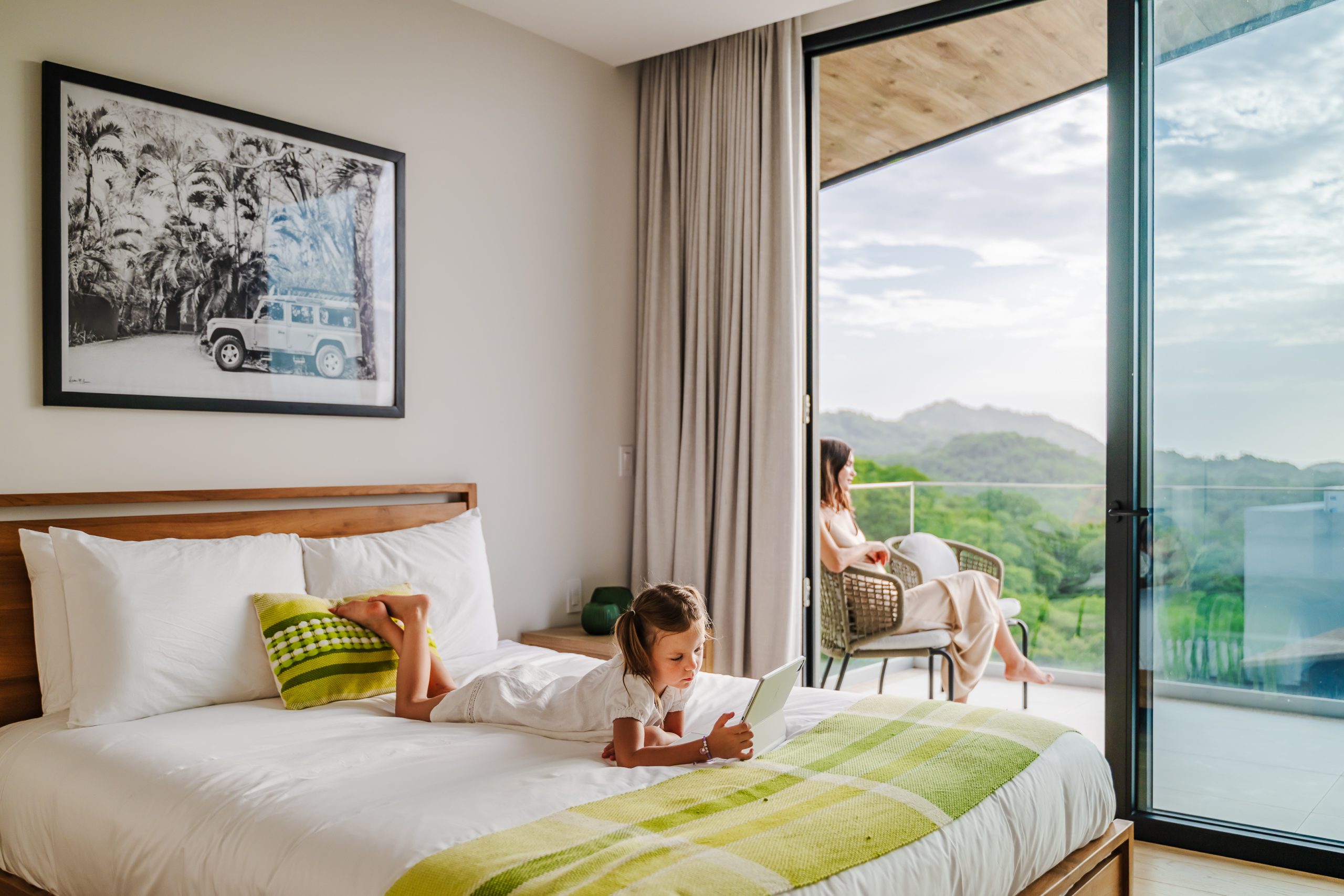 Woman relaxing on the terrace while her child lounges on the bed playing on an iPad inside the Reserva Conchal penthouse, blending comfort and family leisure in a luxury Costa Rican setting.