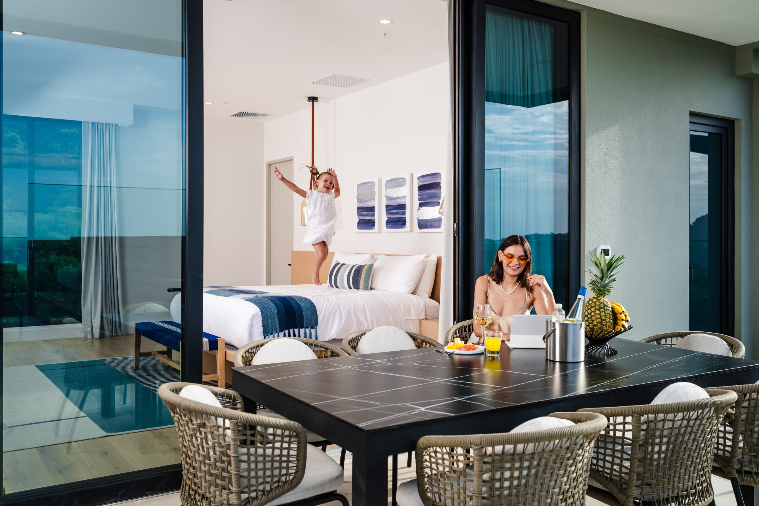 Child playfully jumping on the bed while the mother relaxes on the outdoor terrace of a luxury Reserva Conchal penthouse, overlooking the lush Costa Rican landscape.