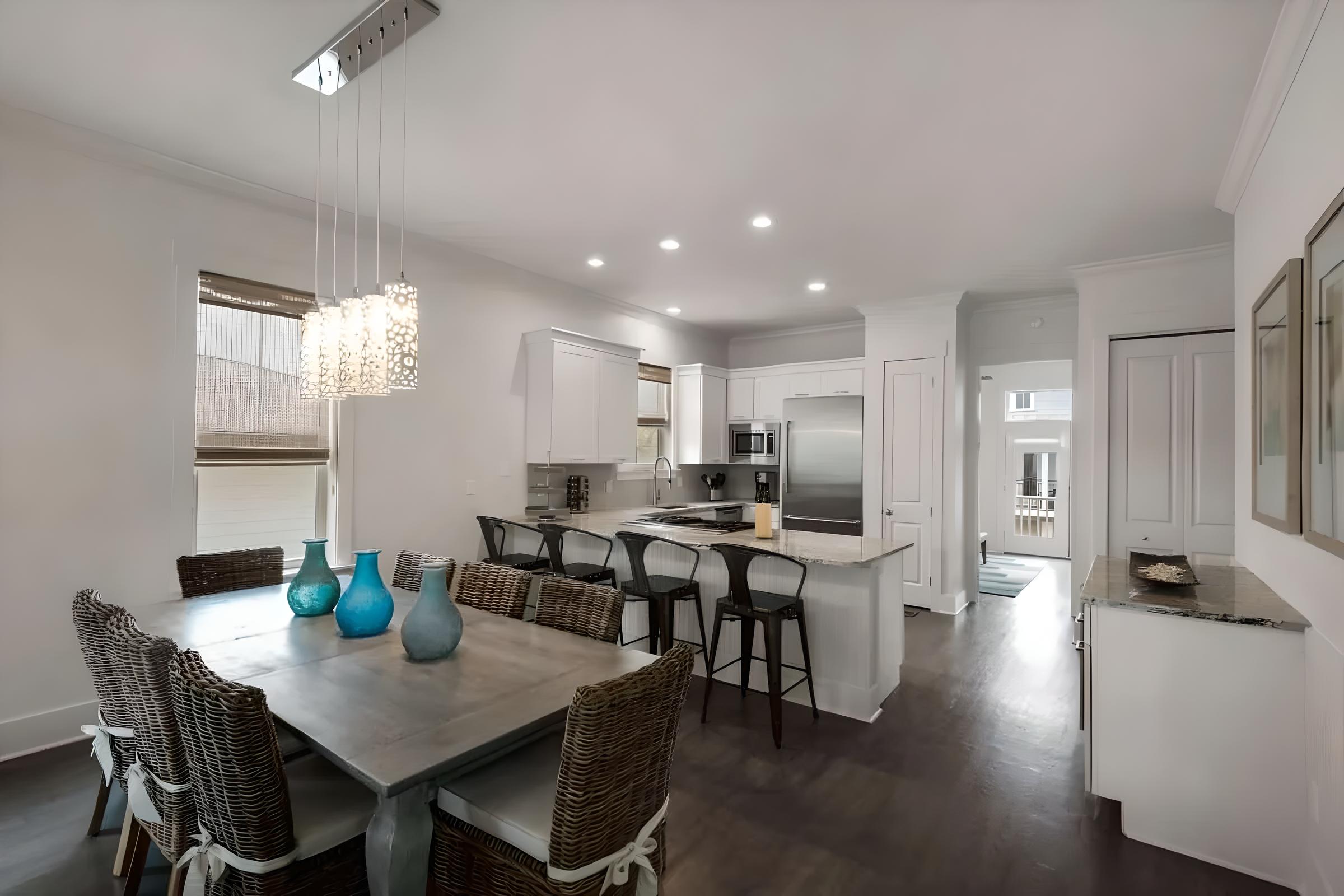 Luxury kitchen and dining area in a modern Seacrest Beach home, featuring a wooden dining table with woven chairs, decorative blue glass vases, stainless steel appliances, and contemporary lighting.