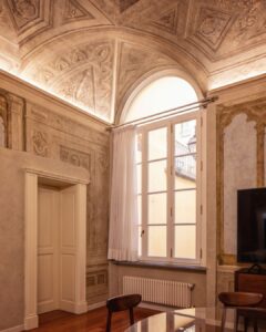 Ornate Renaissance-style ceiling and frescoed walls inside a Florence apartment, featuring tall arched windows, soft white curtains, and warm ambient lighting.