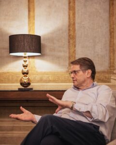 Massimo speaking thoughtfully during an interview in a Florence apartment, seated beside warm lighting and historic frescoed walls.